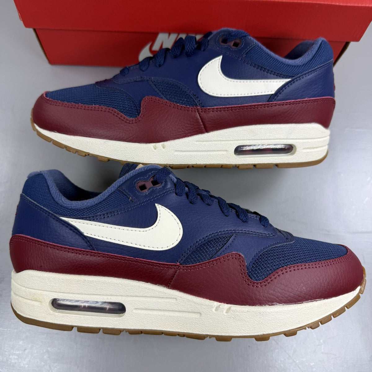 Nike Air Max 1 red trainers with navy accents and gum sole, featuring logo details.