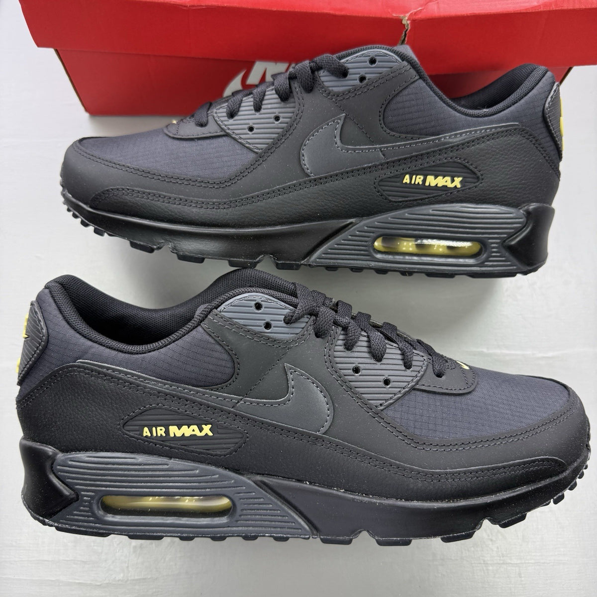 Nike Air Max 90 black trainers with buff gold and anthracite grey accents.