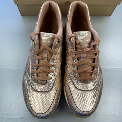 Nike Air Max 1 white trainers with bronze snake print and metallic accents.