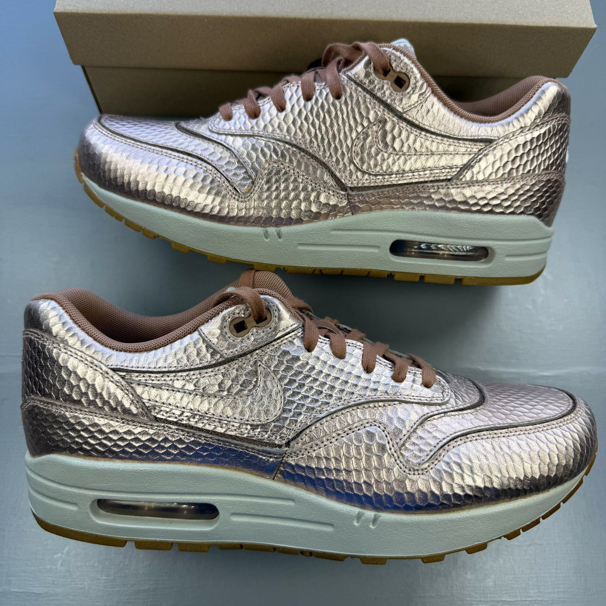 Nike Air Max 1 white trainers with bronze snake print and metallic accents.