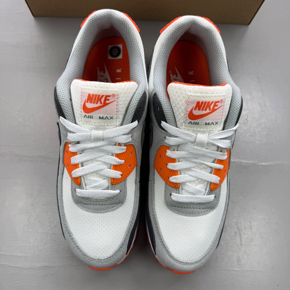 Nike Air Max 90 white trainers with safety orange accents and grey detailing.