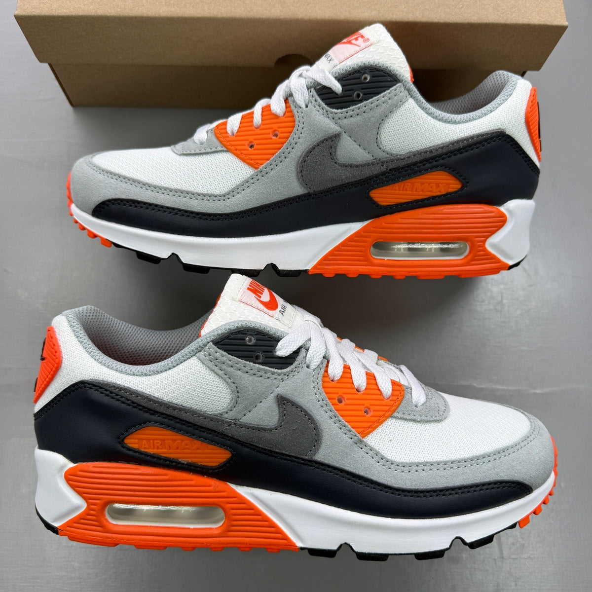 Nike Air Max 90 white trainers with safety orange accents and grey detailing.