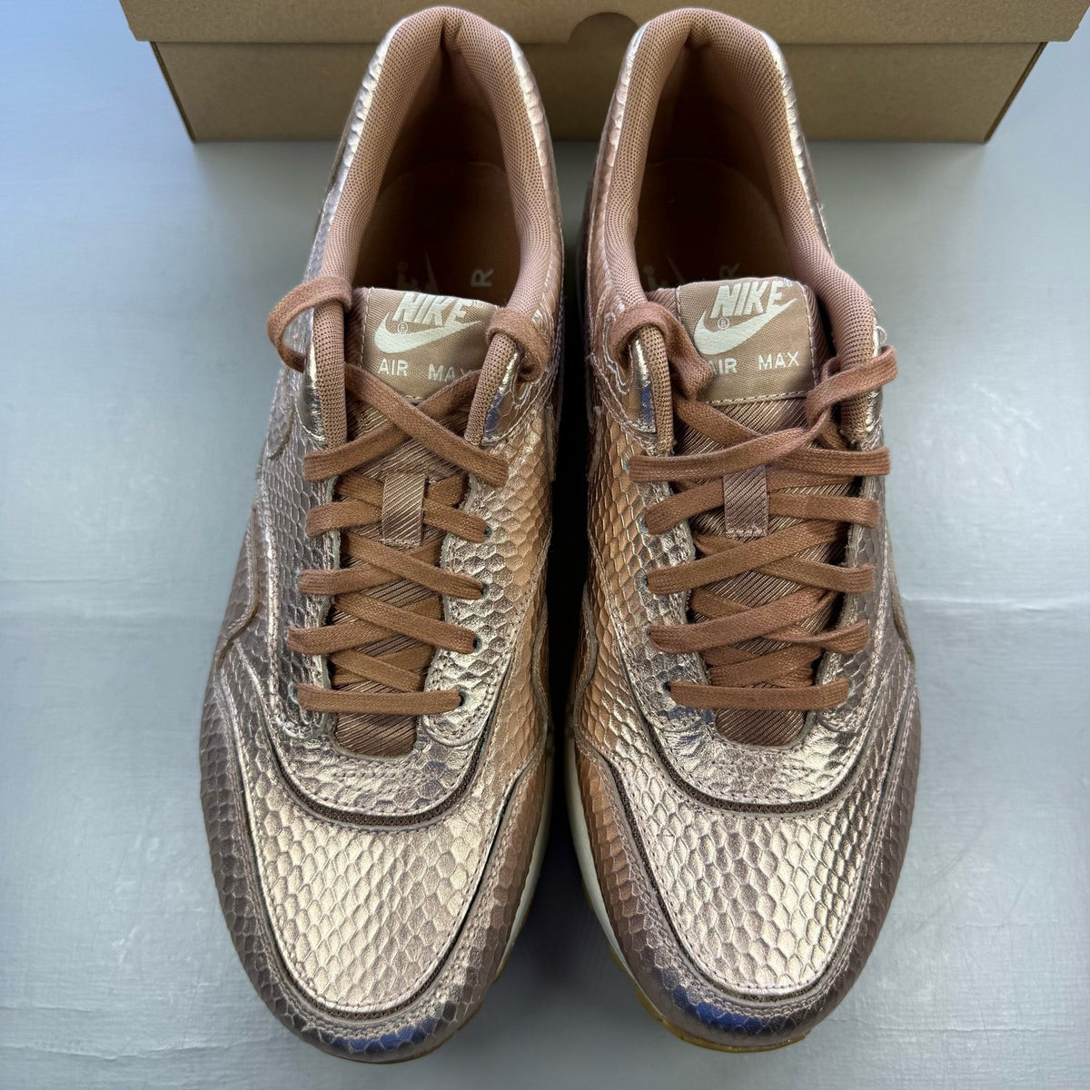 Nike Air Max 1 white trainers with bronze snake print and metallic accents.