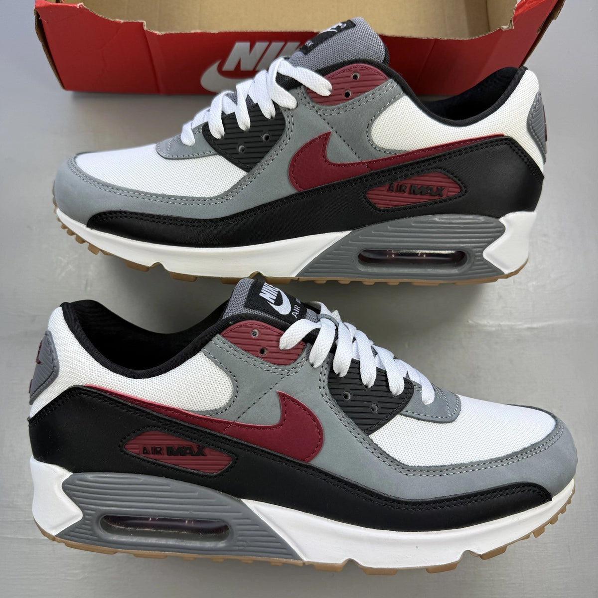 Nike Air Max 90 grey trainers with red accents and gum sole, featuring logo details.